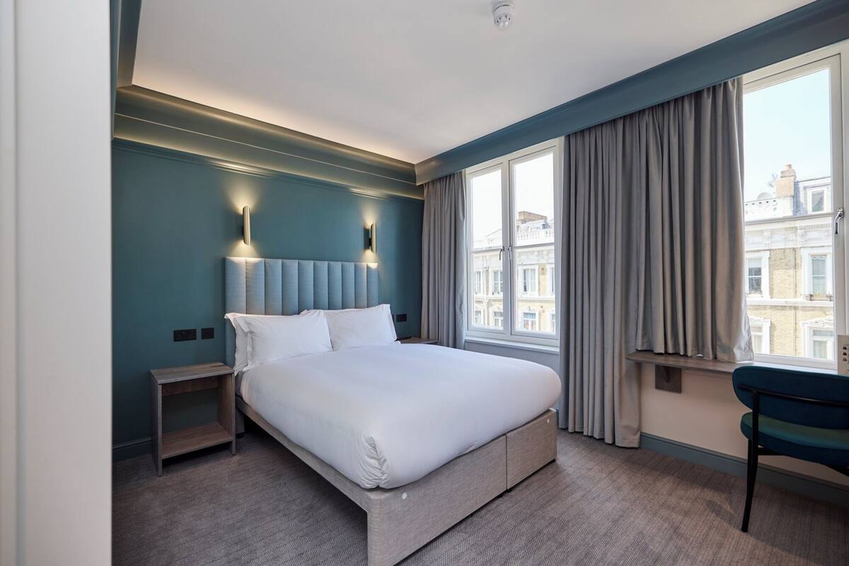 A hotel room with a large bed, two bedside tables, a chair, a window with curtains, and a view of buildings outside.