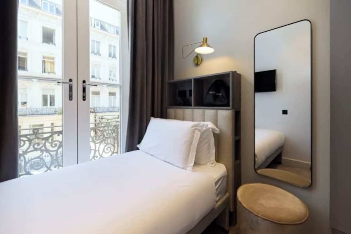A hotel room with a bed, a bedside table, a mirror, and a window with a view of buildings.