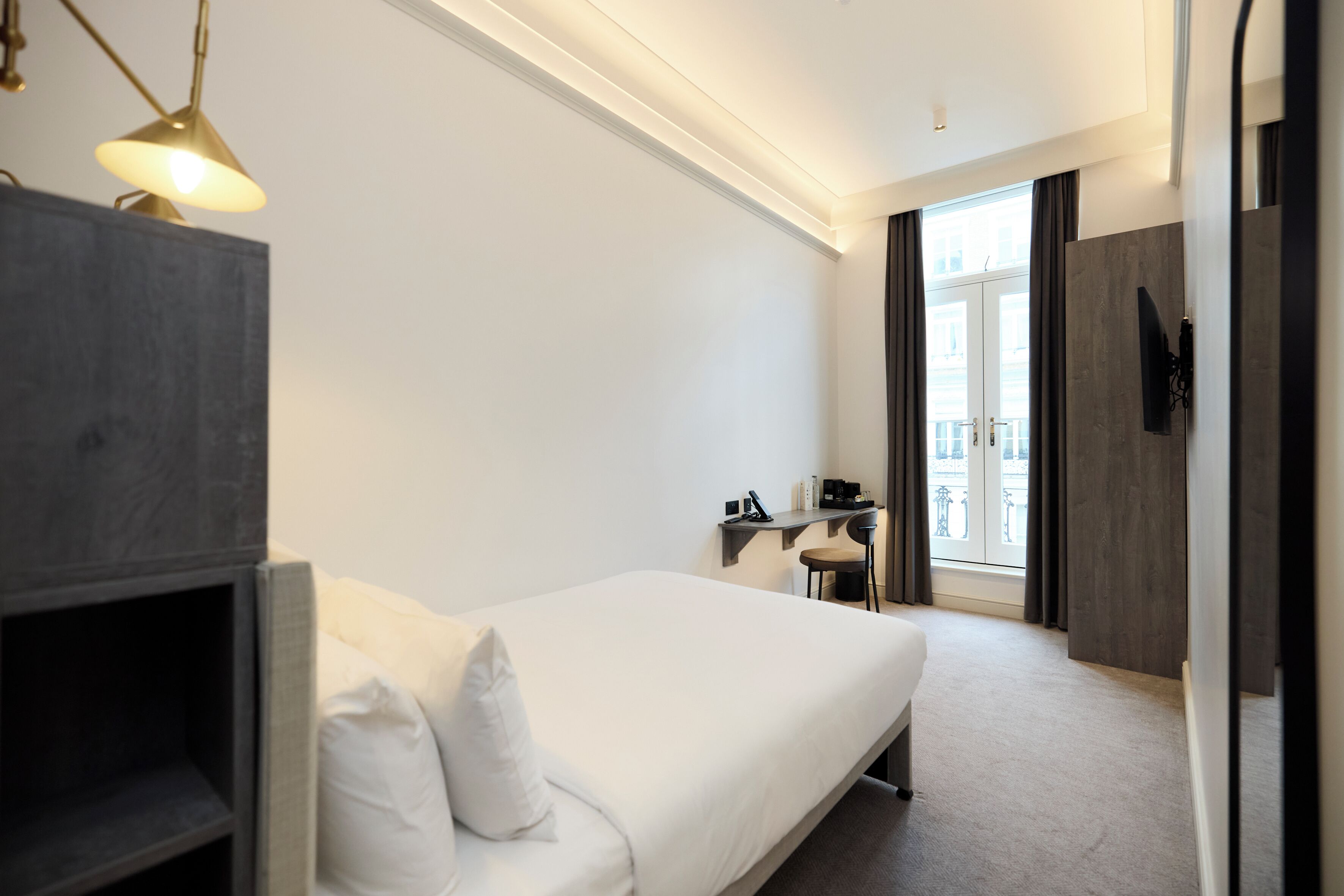 A modern hotel room with a large bed, a desk with a chair, a flat-screen TV mounted on the wall, and a window with curtains.