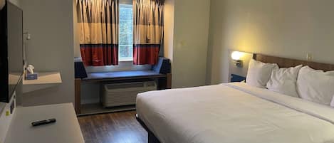A hotel room with a bed, a desk, a chair, a window with curtains, and a nightstand with a lamp.