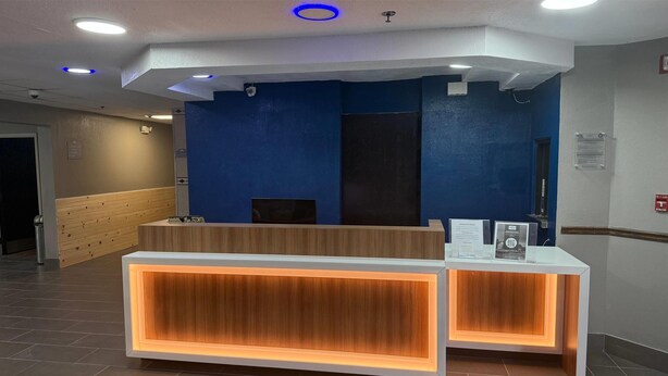 A reception desk with a wooden finish and illuminated panels, a blue wall, and a tiled floor. - Microtel Inn by Wyndham Atlanta Airport (College Park)
