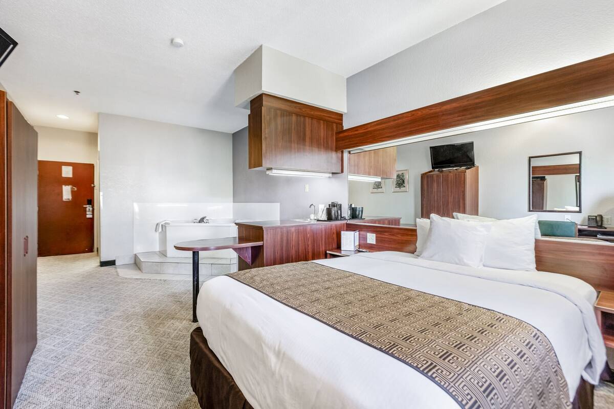 Quarto de hotel com cama, escrivaninha, espelho, televisão e banheiro com pia e chuveiro.