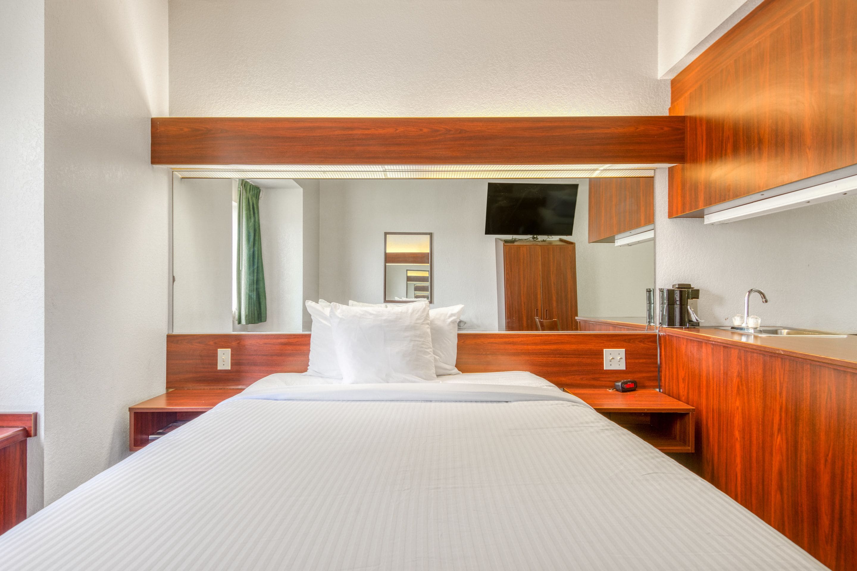 A hotel room with a large bed, wooden headboard, a small desk with a lamp, a mirror, and a television mounted on the wall.