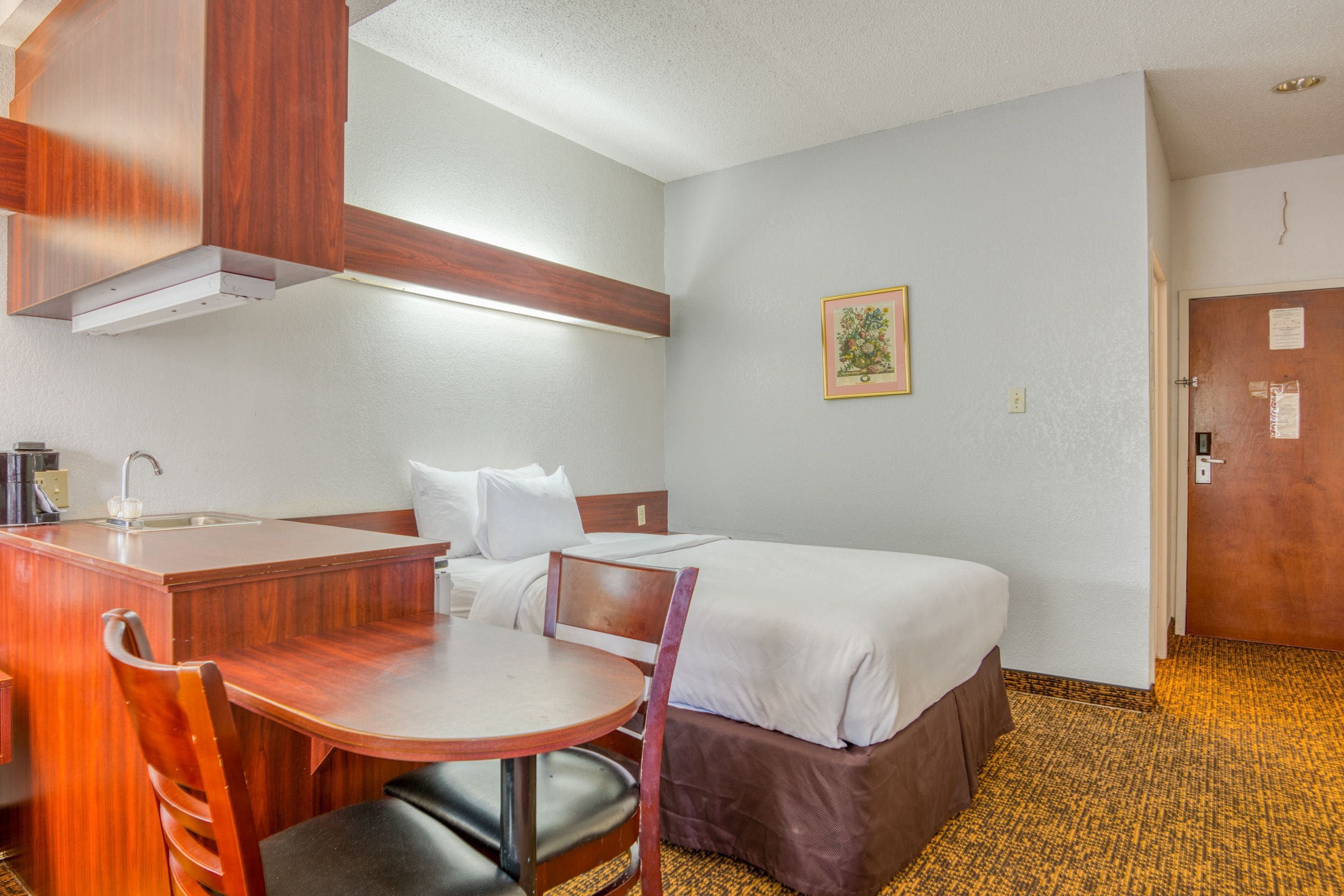 Una camera d'albergo compatta con angolo cottura, un letto singolo, un tavolino con una sedia e una porta.