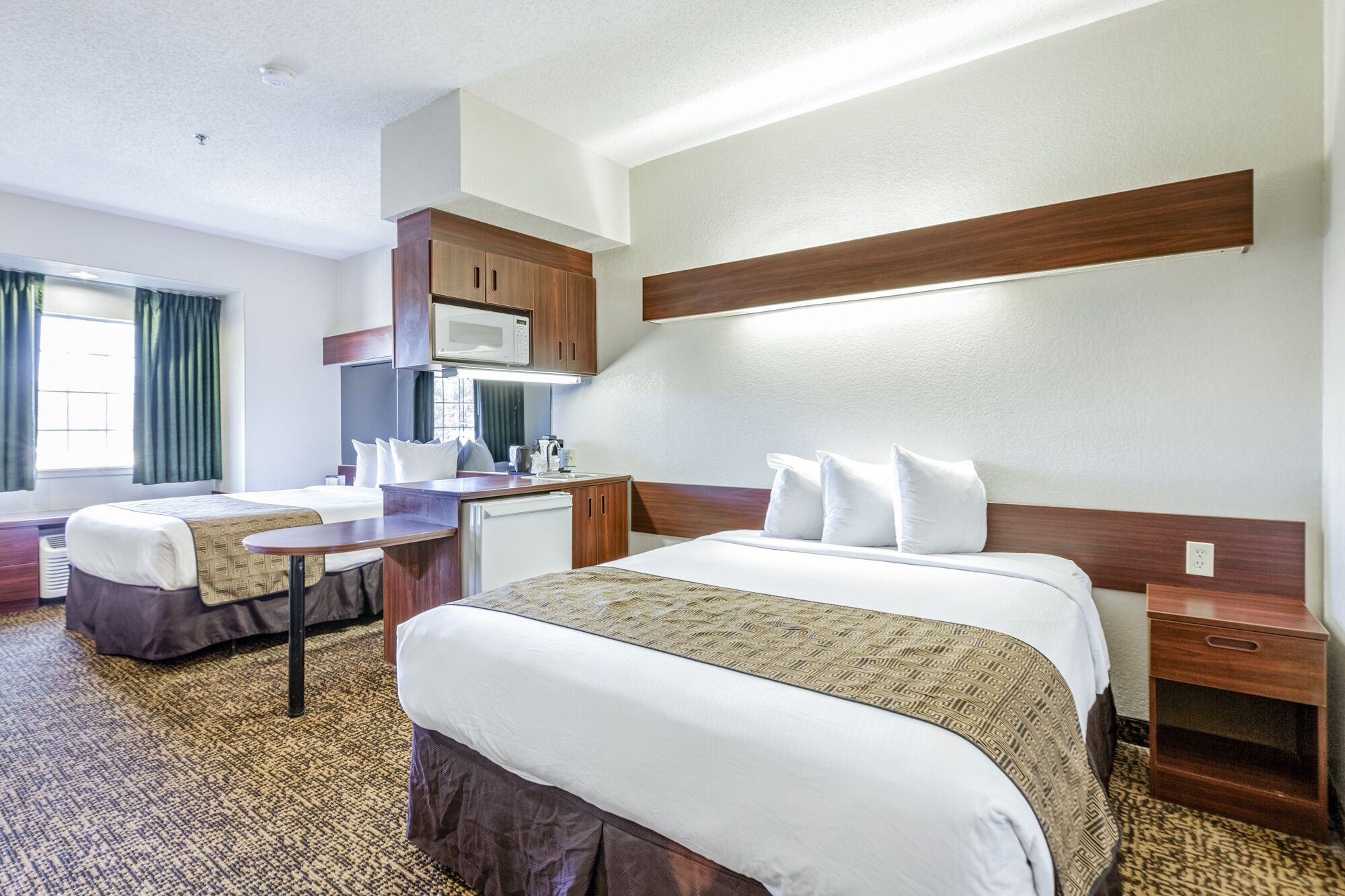 A hotel room with two beds, a kitchenette, and a small desk.