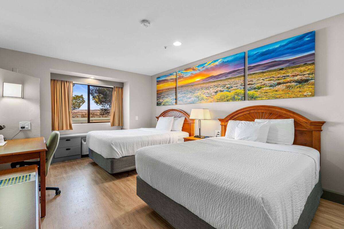 Habitación de hotel con dos camas, un escritorio, una silla, una ventana con cortinas y un cuadro grande con un paisaje.