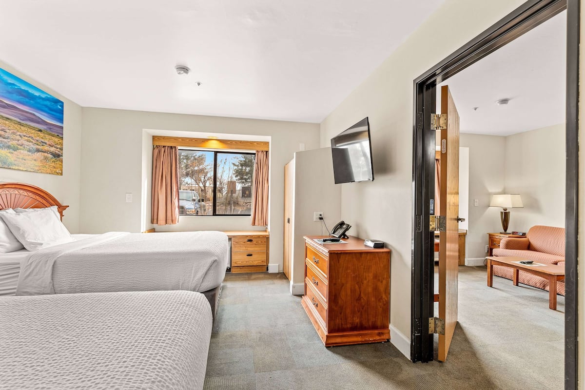 Habitación de hotel con dos camas, un aparador de madera, un escritorio con silla, un televisor montado en la pared y una ventana con cortinas.