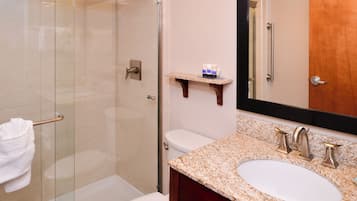 A bathroom with a glass-enclosed shower, a white toilet, a granite countertop sink, and a wooden shelf with toiletries.