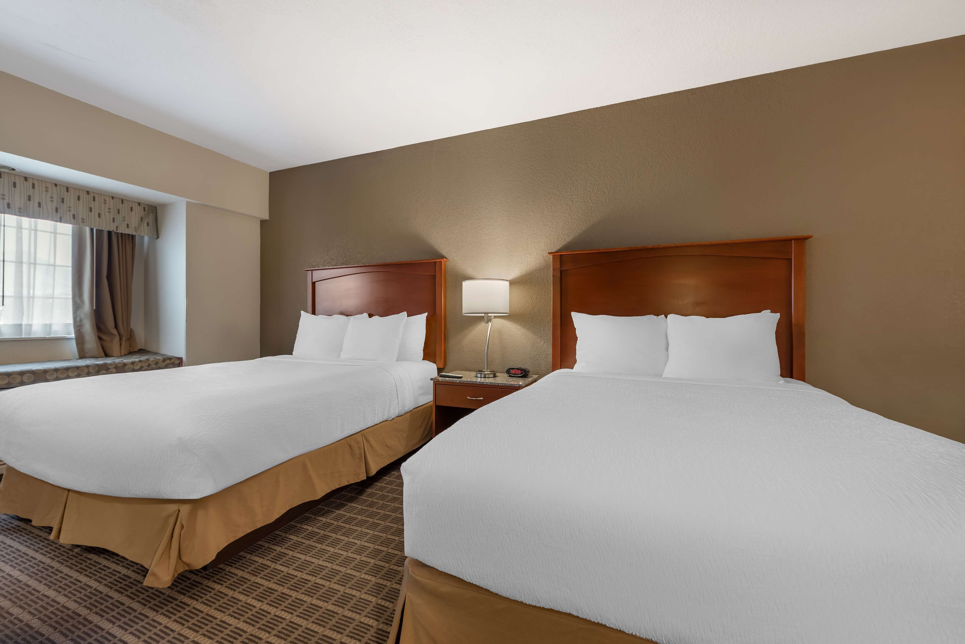 Quarto de hotel com duas camas, uma mesa de cabeceira, uma luminária e uma janela com cortinas.