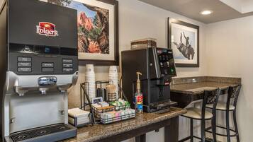 A coffee station with a Folgers coffee maker, a hot water dispenser, and a refrigerator. There are bar stools and a framed picture on the wall.