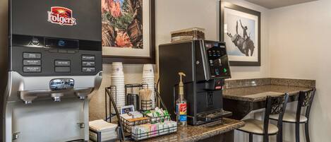 A coffee station with a Folgers coffee maker, a hot water dispenser, and a refrigerator. There are bar stools and a framed picture on the wall.