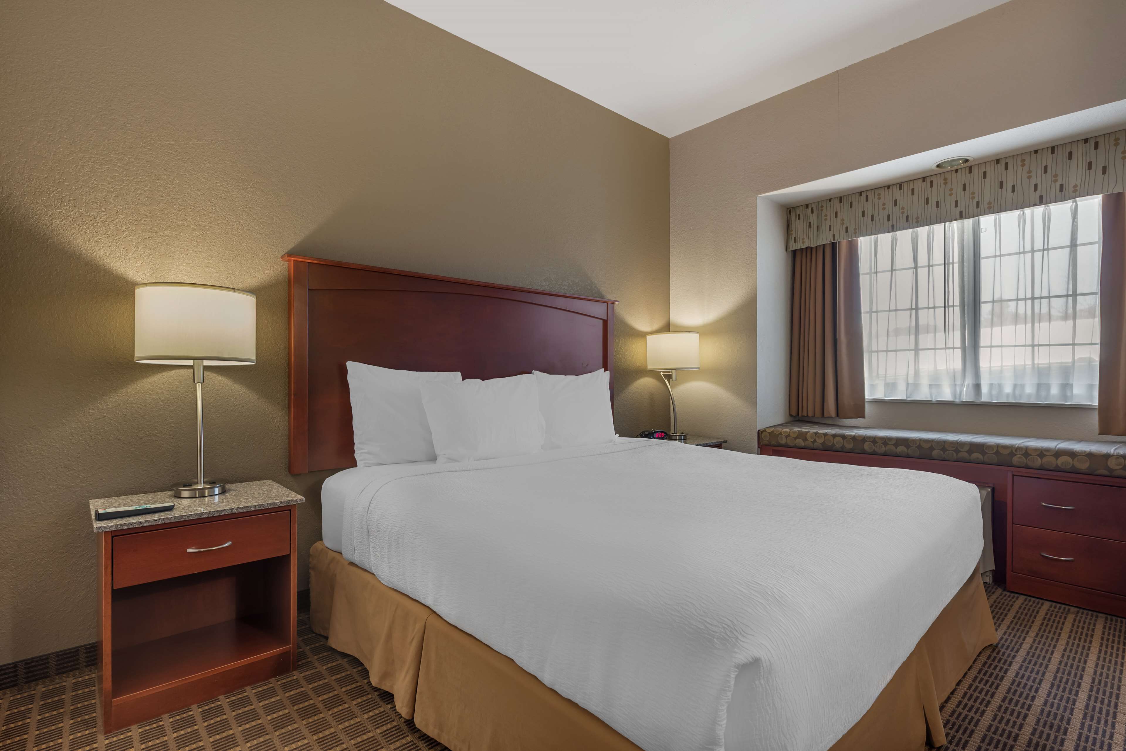 A hotel room with a large bed, two bedside tables with lamps, a window with curtains, and a carpeted floor.