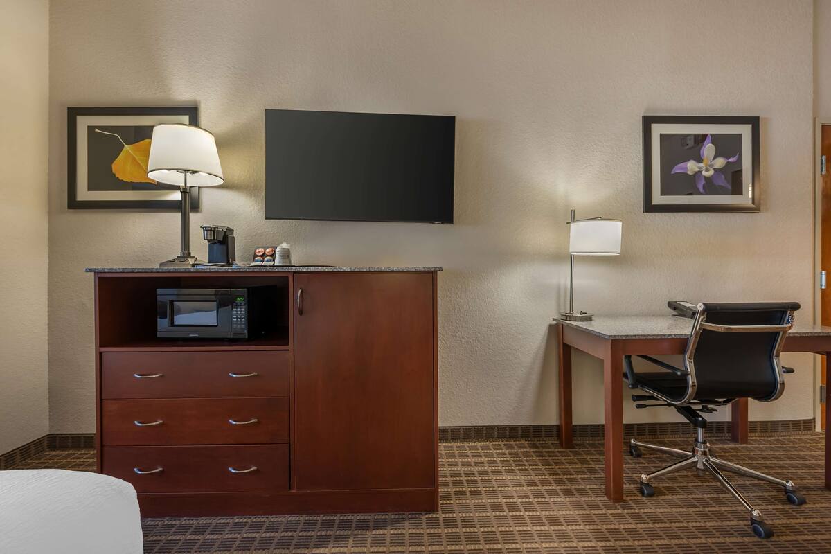 A hotel room with a wooden desk, a black office chair, a flat-screen TV, a microwave, a lamp, and a minibar.