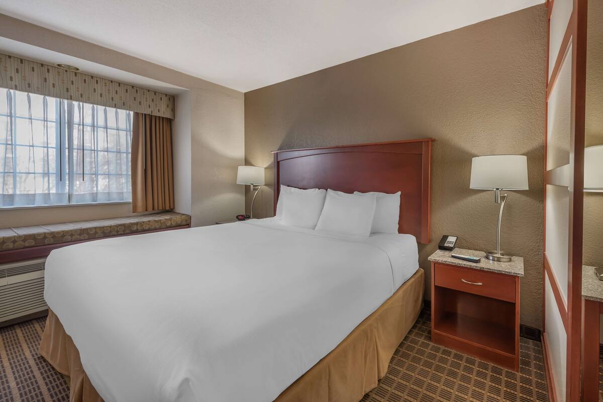 A hotel room with a large bed, two bedside tables with lamps, a window with curtains, and a built-in bench under the window.