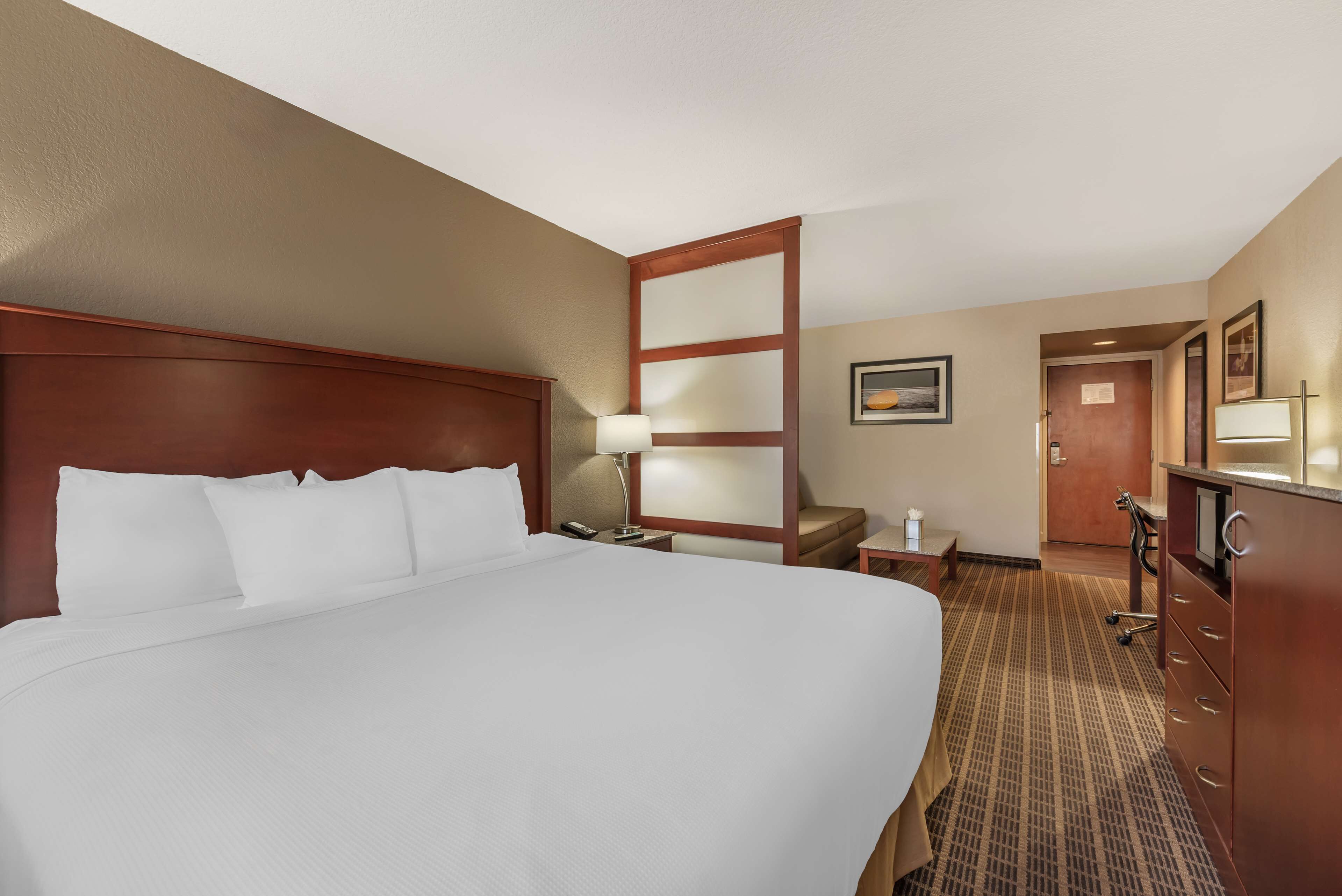 Una habitación de hotel con una cama grande, un cabecero de madera, un escritorio con lámpara, una silla, un aparador y una puerta que da a otra habitación.