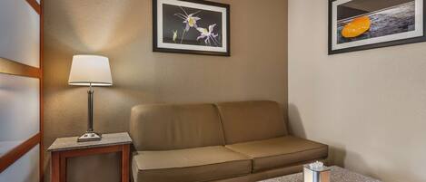 A hotel room with a beige sofa, a small table with a lamp, and two framed pictures on the wall.