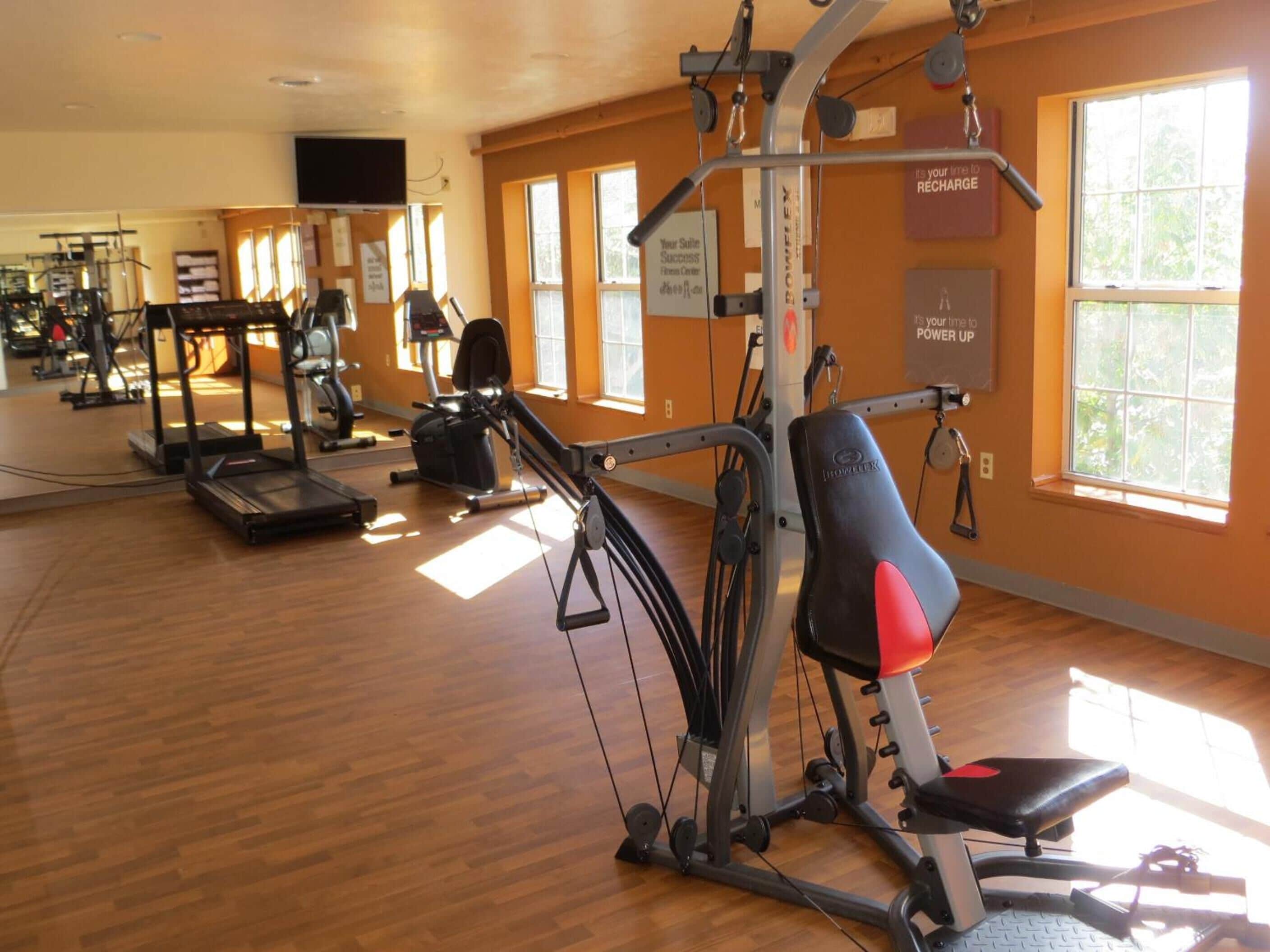 A gym with various exercise equipment, including treadmills and weight machines, in a room with large windows and wooden floors.
