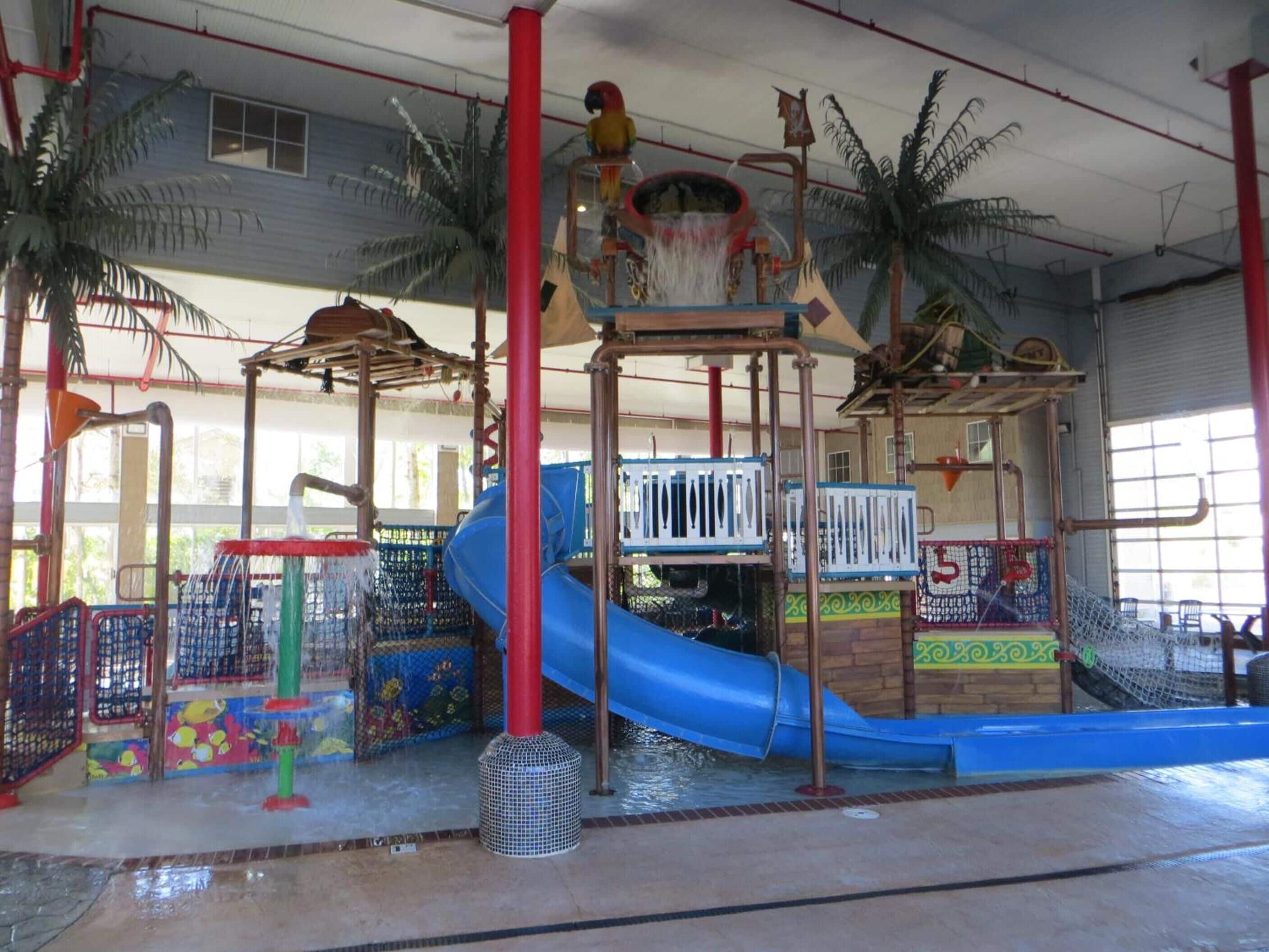 Un área de juegos interior con un tobogán azul, estructuras para escalar y una zona de juegos acuáticos.