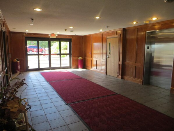 A hotel lobby with a red carpet, tiled floor, wooden paneling, an elevator, and a glass door leading outside.