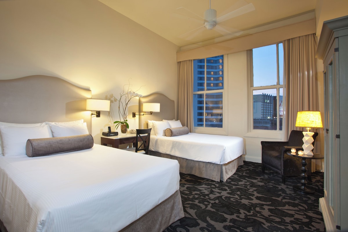 A hotel room with two beds, a desk, a chair, a lamp, and a view of buildings through the window.