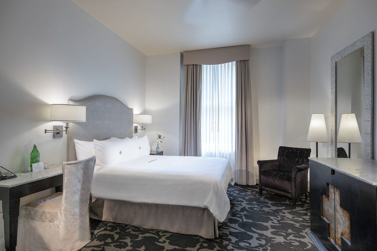 Une chambre d’hôtel avec un grand lit, un bureau avec une lampe, une chaise, une commode et un grand miroir.