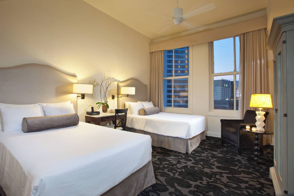 A hotel room with two beds, a desk, a chair, a lamp, and a view of buildings through the window.