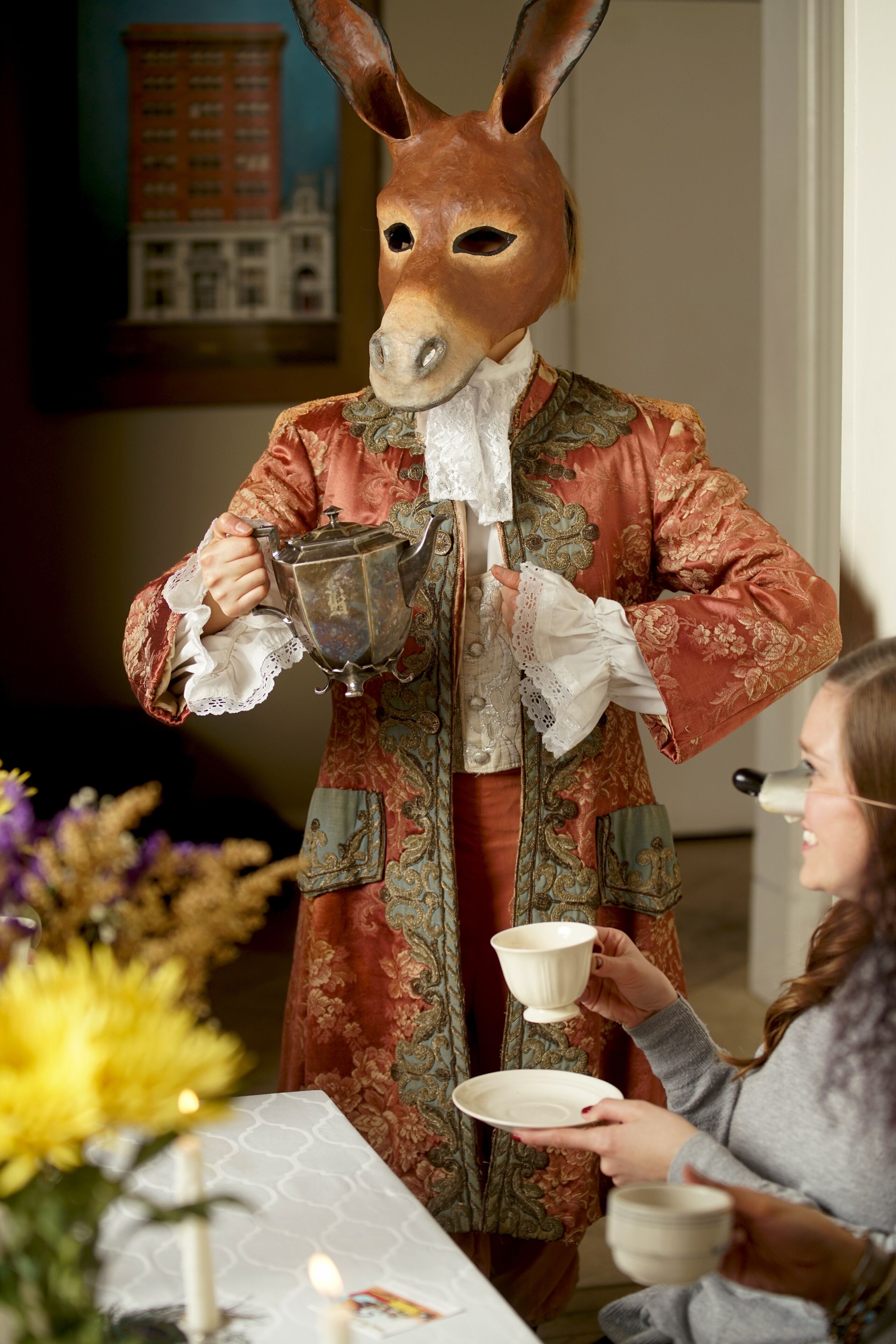 Eine Person in einem Hirschkostüm hält eine Teekanne und eine Tasse, stehend an einem Tisch mit einer weiteren Person.