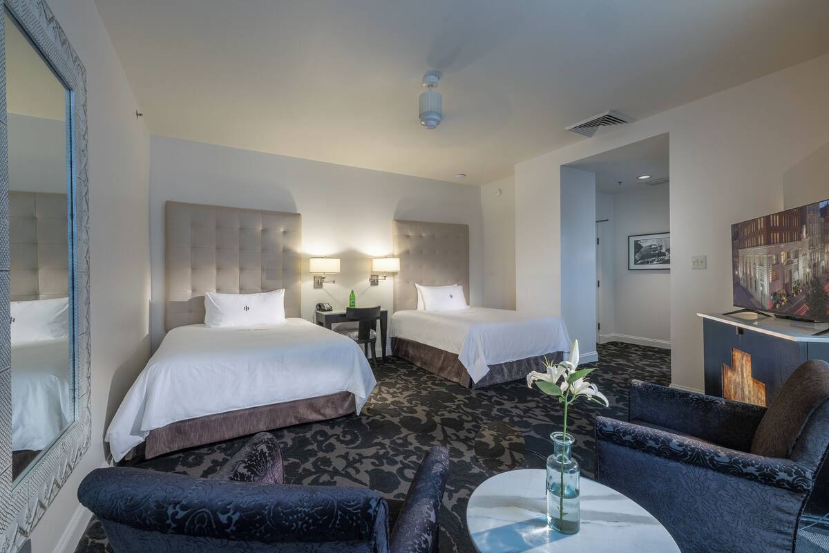 Une chambre d’hôtel avec deux lits, un bureau, une chaise, une petite table avec un vase et un tableau au mur.