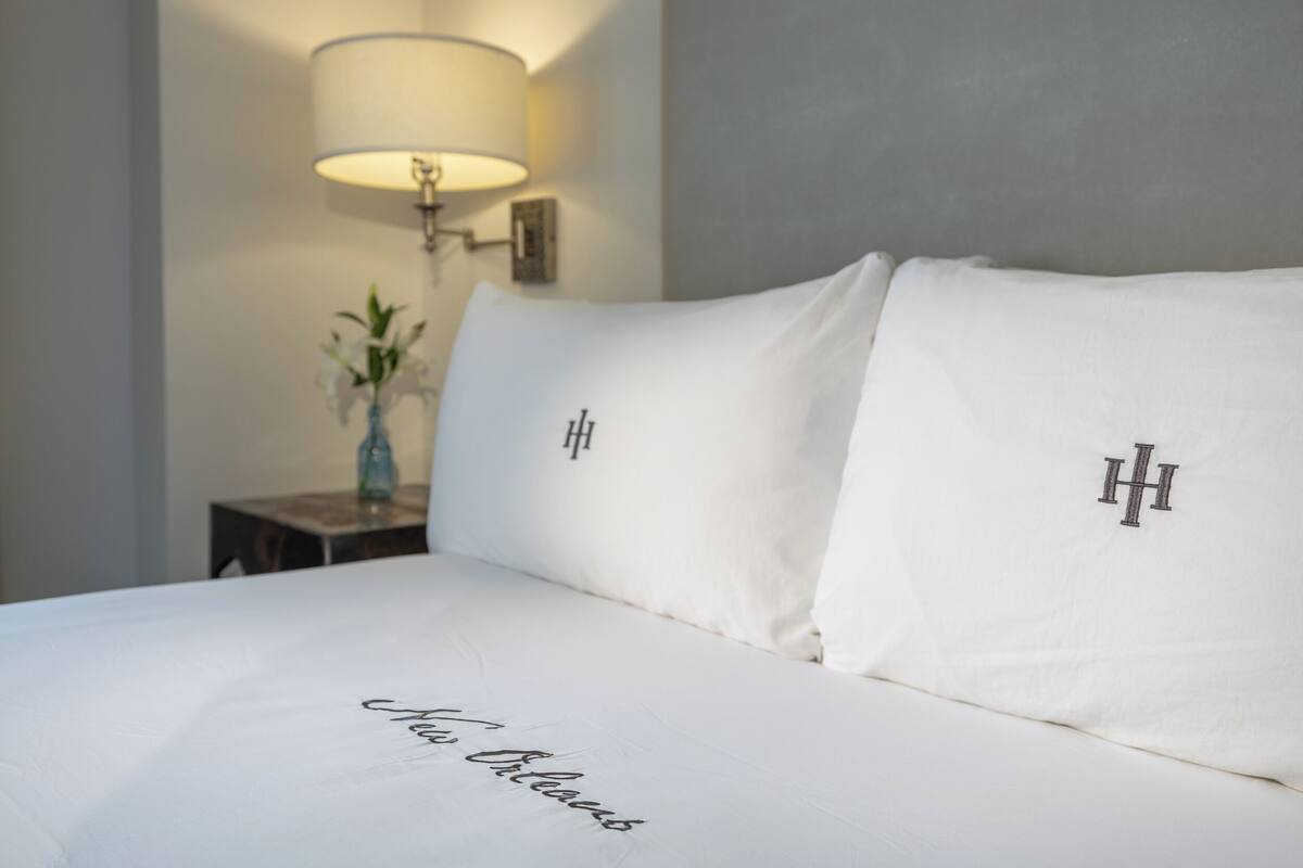 Un lit bien fait, avec du linge de lit blanc et des oreillers ornés de monogrammes brodés. On aperçoit une table de chevet avec une lampe et un vase de fleurs.