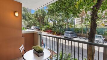 A balcony with a table and chair, a potted plant, and a view of a street with cars and trees.