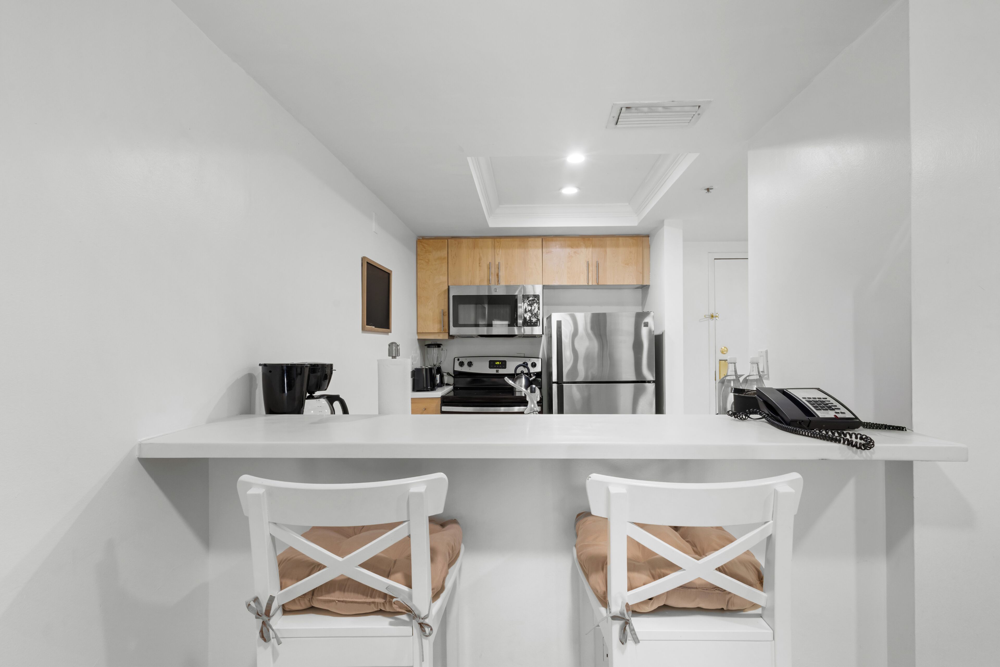 A modern kitchen with white cabinetry, a microwave, a refrigerator, and a coffee machine. There are two white chairs with brown cushions.
