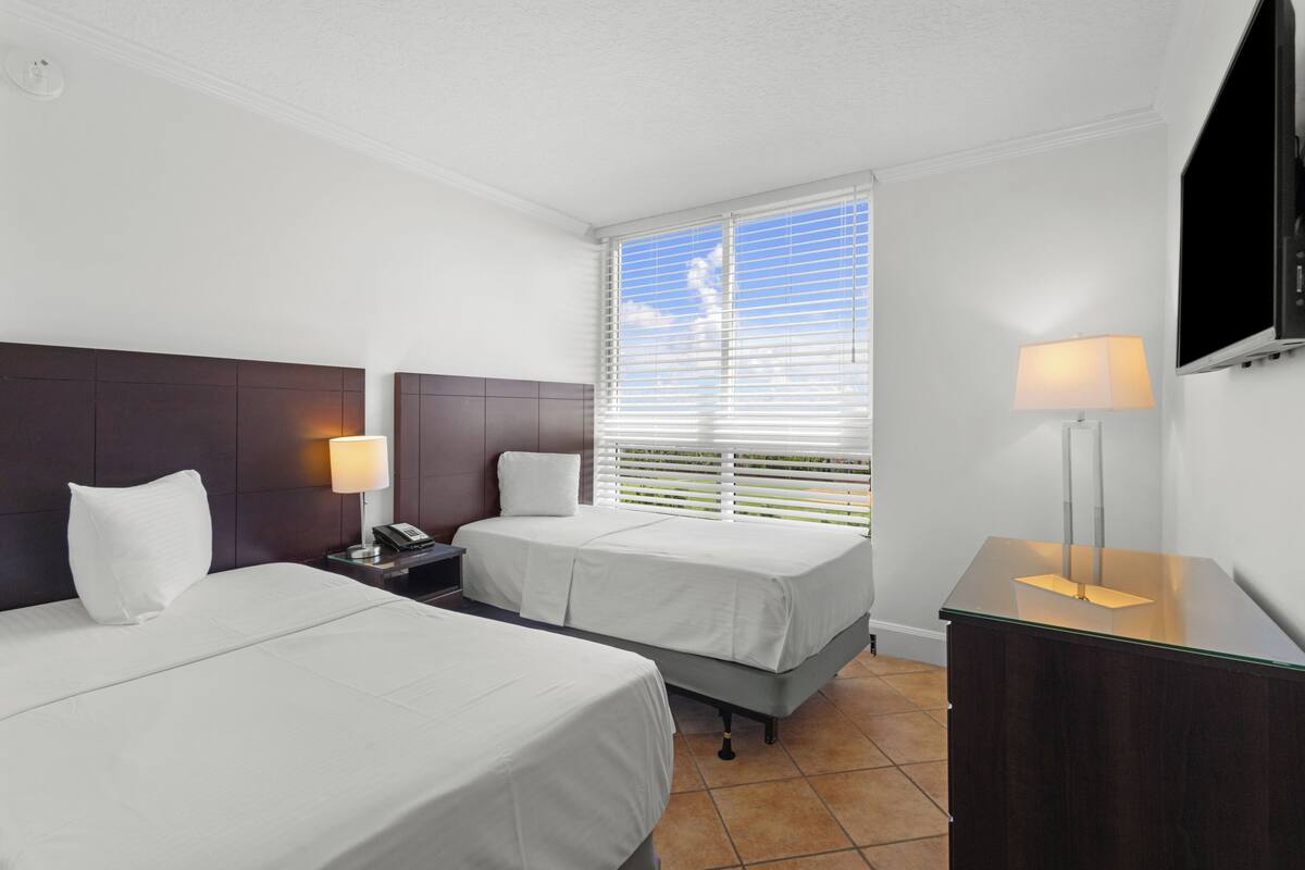 Habitación de hotel con dos camas, una mesita de noche con lámpara, un escritorio con lámpara, televisión y ventana con persianas.