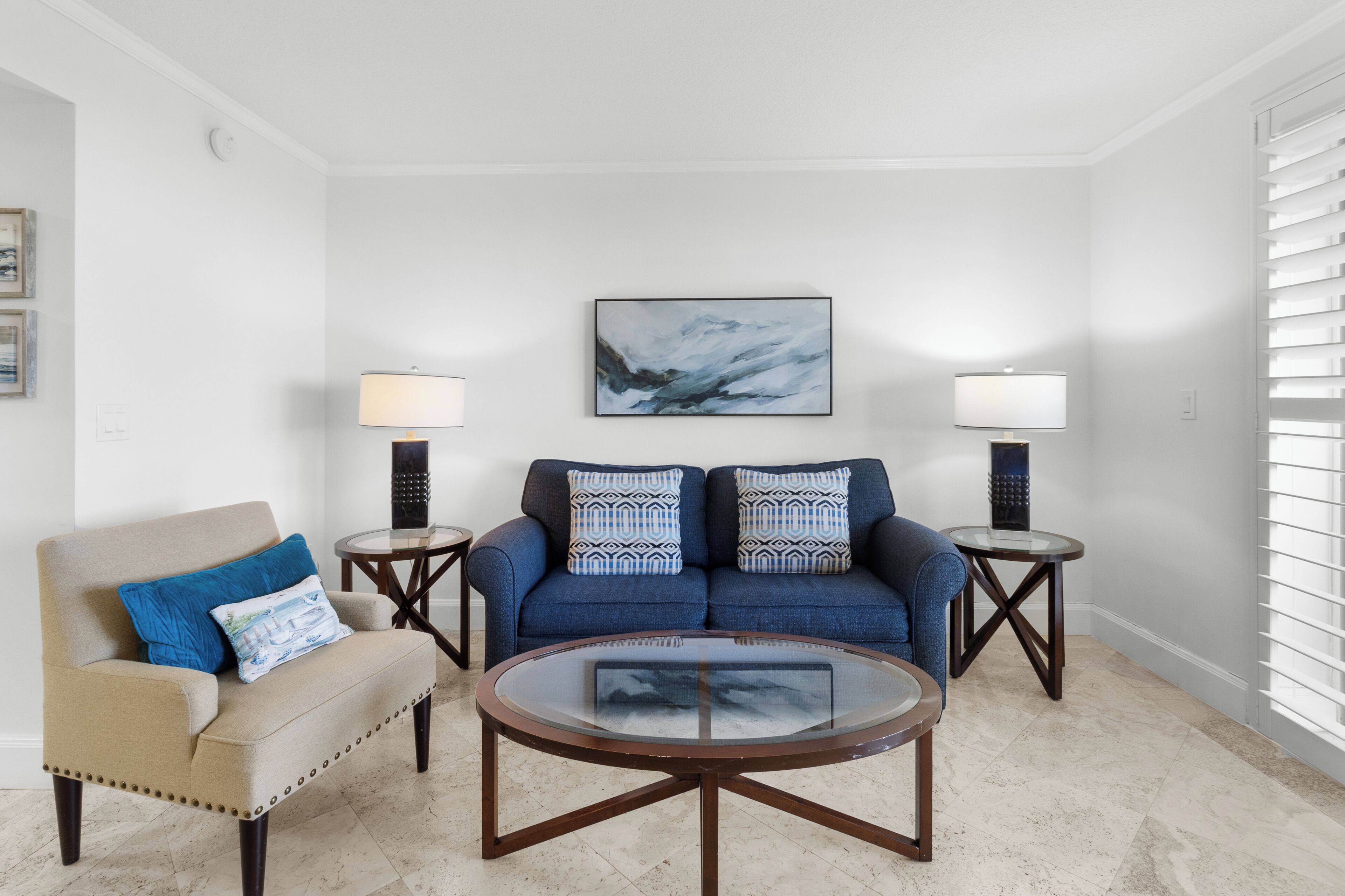 A living room with a beige armchair, a blue sofa, two side tables with lamps, a glass coffee table, and a painting on the wall.