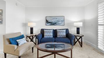 A living room with a beige armchair, a blue sofa, two side tables with lamps, a glass coffee table, and a painting on the wall.