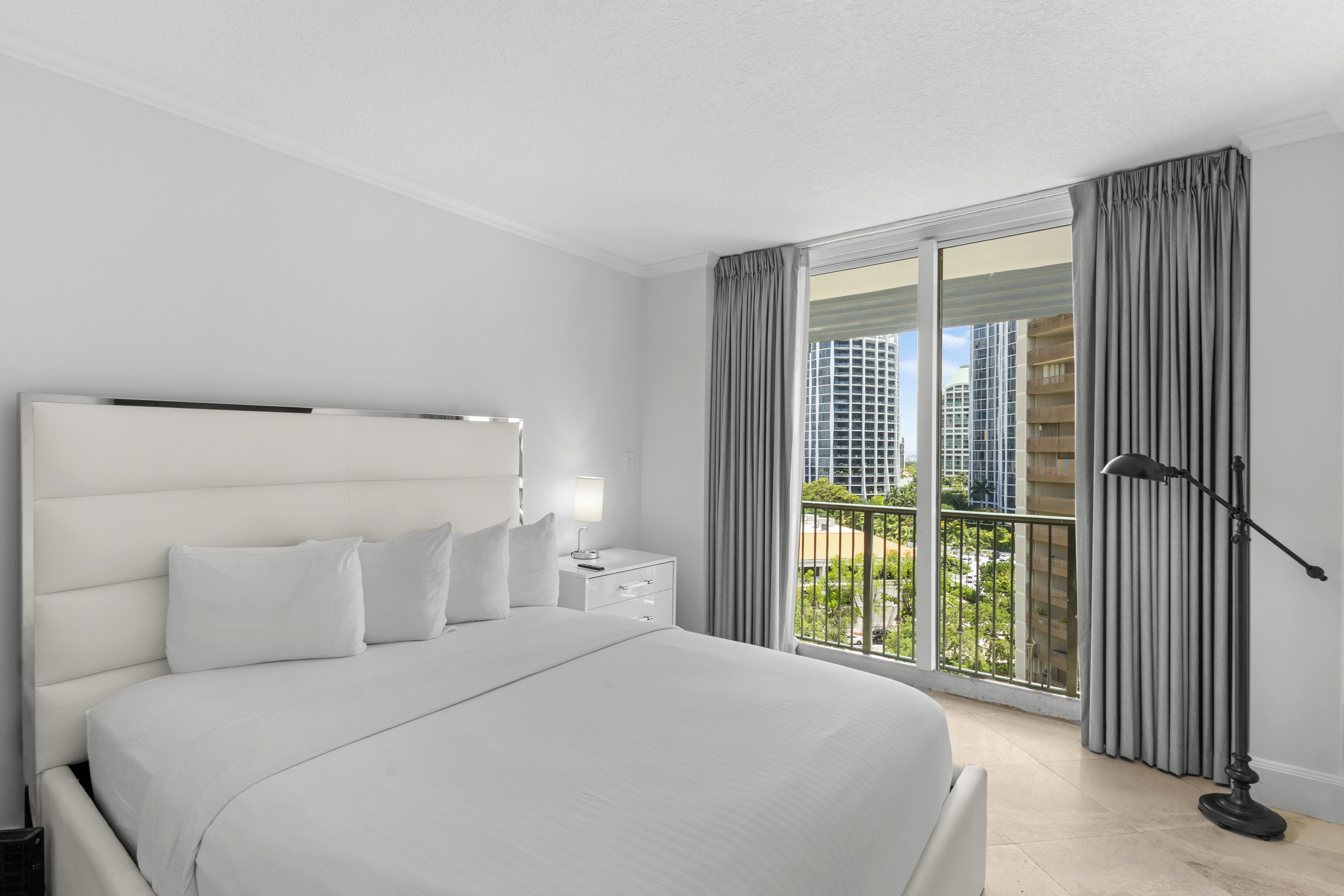 Una habitación de hotel moderna con una cama grande, una mesita de noche, una lámpara y vistas a la ciudad desde un balcón.