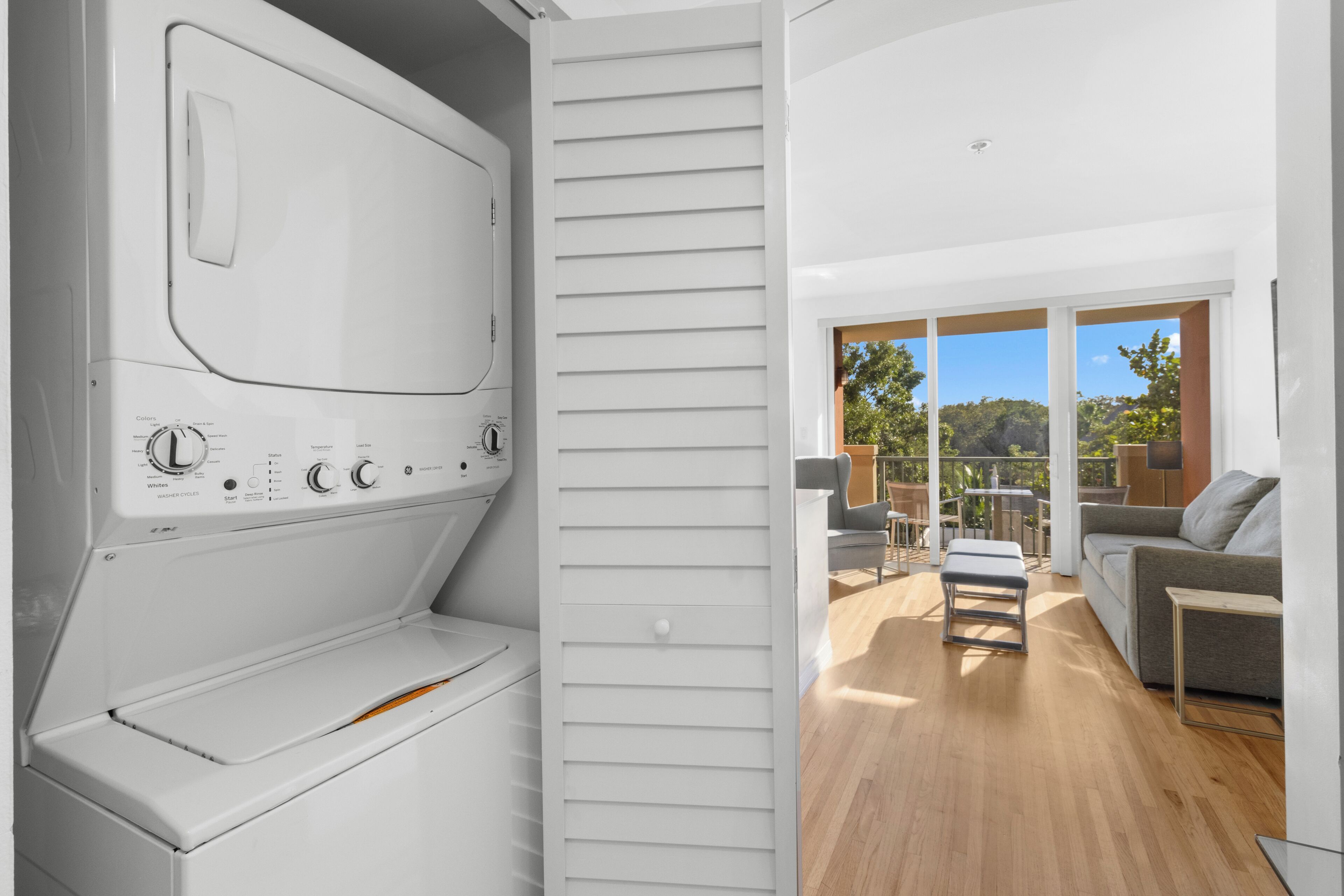 Une buanderie moderne équipée d’un lave-linge et d’un sèche-linge intégrés, avec une porte coulissante donnant sur un balcon aménagé avec du mobilier d’extérieur.