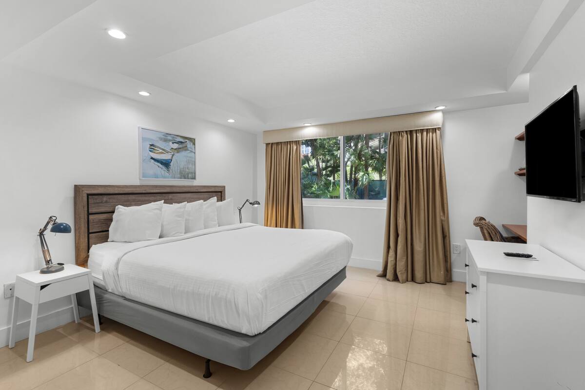 A modern hotel room with a large bed, a desk, a TV, and a window with curtains.