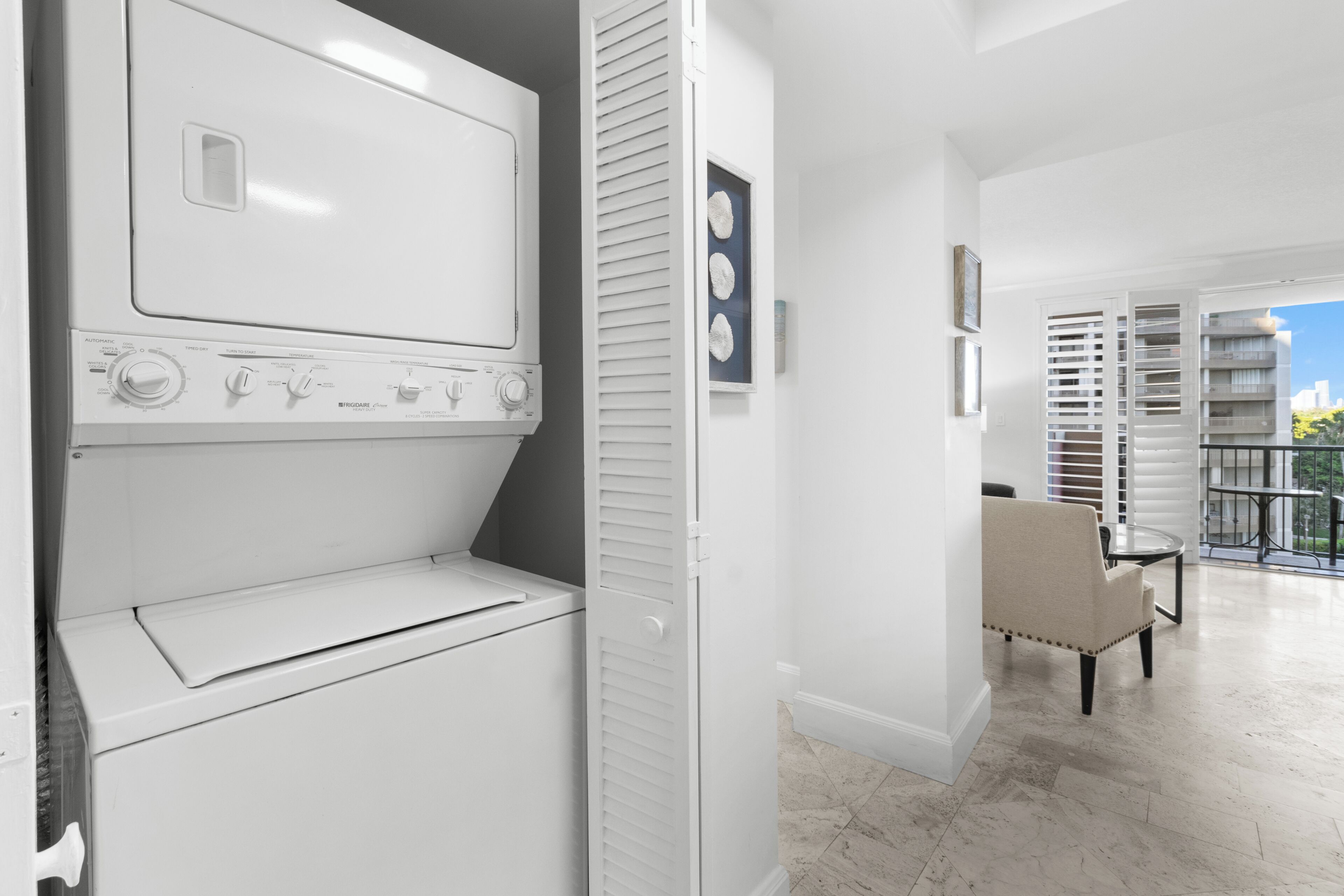 A laundry room with stacked washers and dryers, a chair, and a view of a balcony with outdoor furniture.