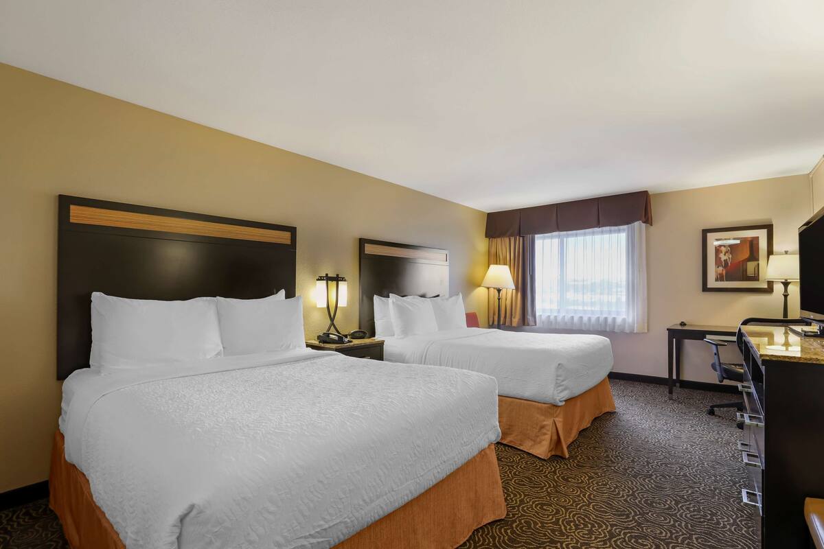 A hotel room with two beds, a desk, a chair, a lamp, and a window with curtains.