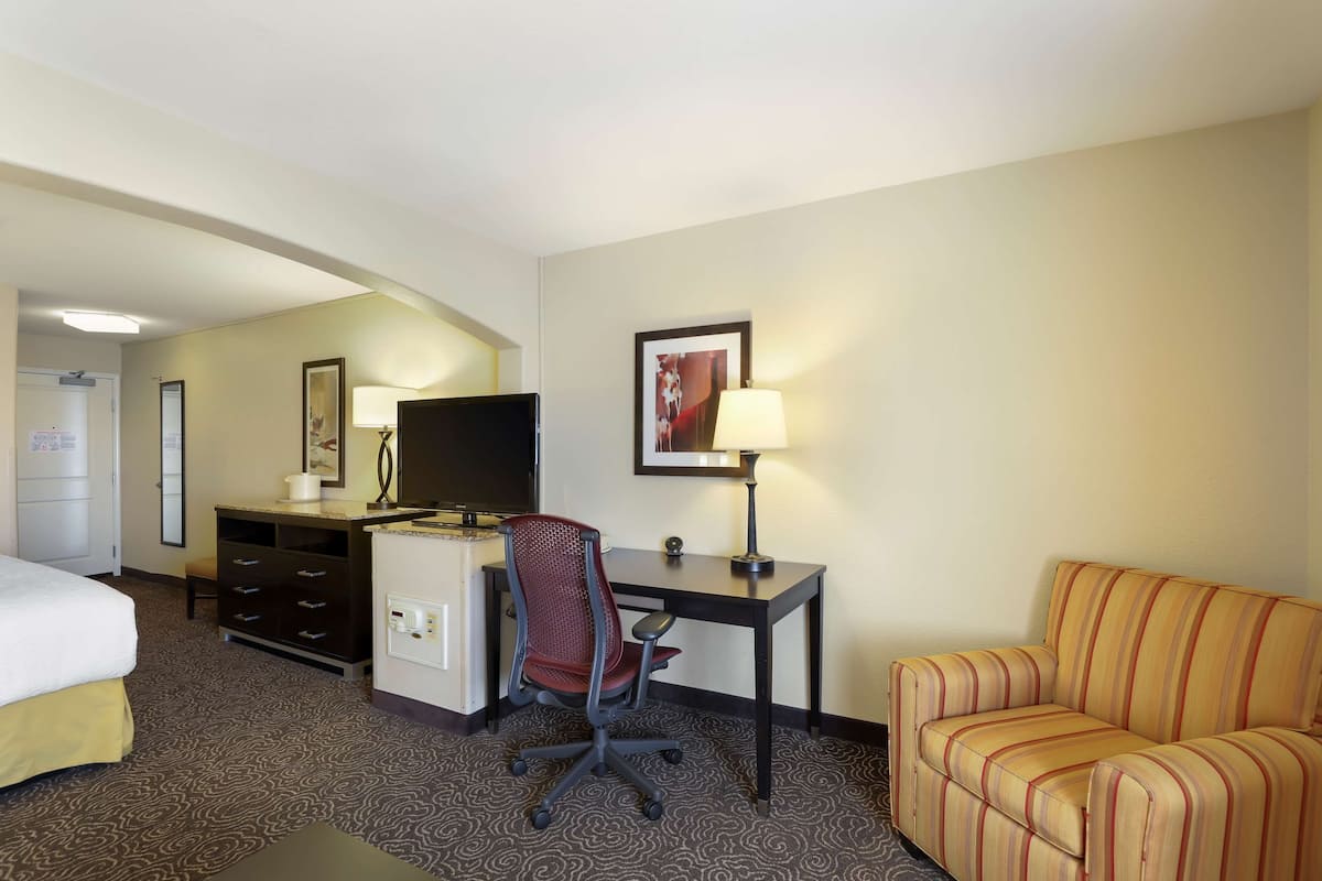 A hotel room with a bed, a desk with a computer, a TV, a chair, a sofa, and a lamp.