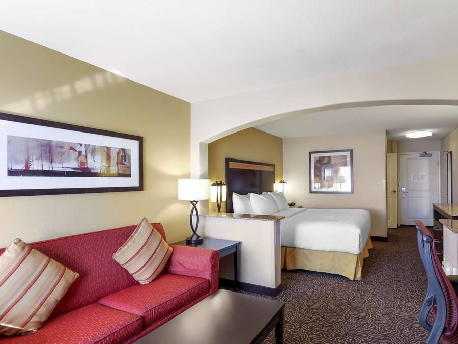 Quarto de hotel com um sofá vermelho, uma cama com roupa de cama branca, uma mesa de trabalho com abajur e uma televisão.