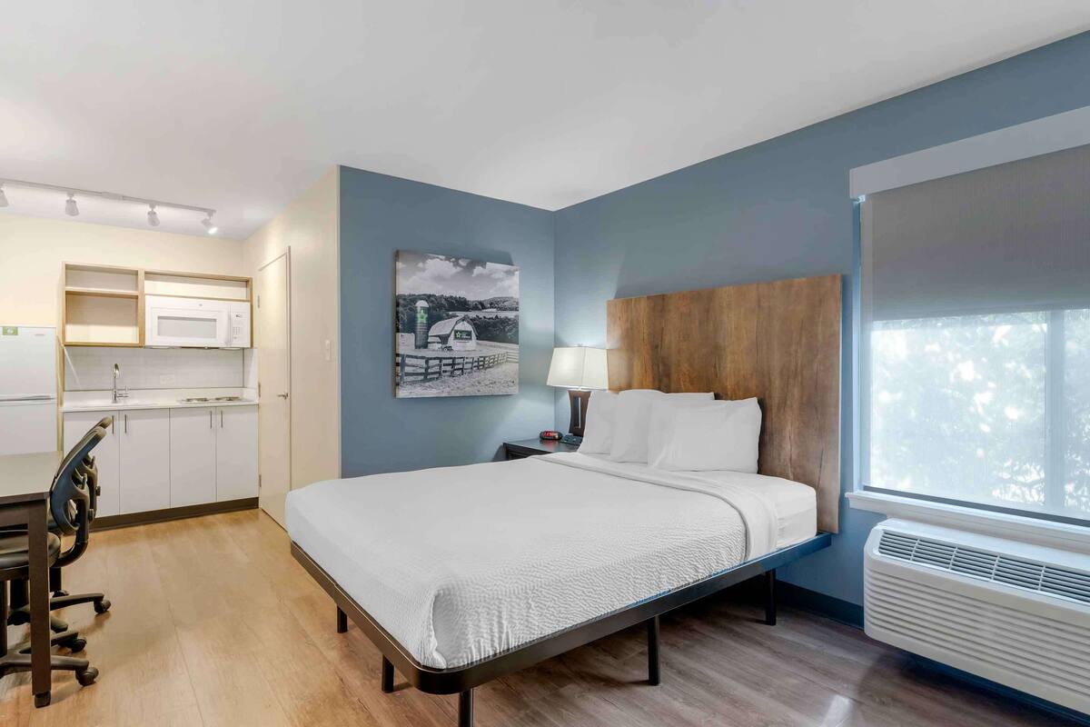 A hotel room with a bed, a desk, a microwave, and a window with blinds.