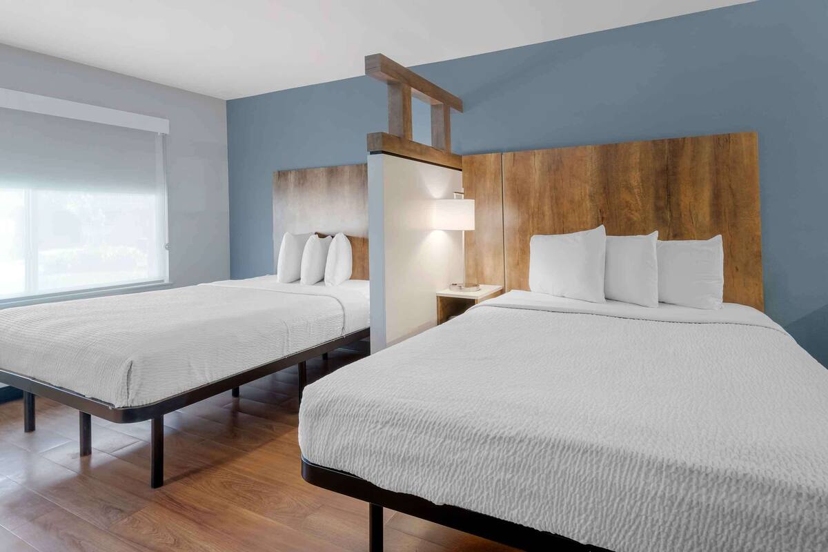 A hotel room with two beds, a wooden headboard, a nightstand with a lamp, and a window with blinds.