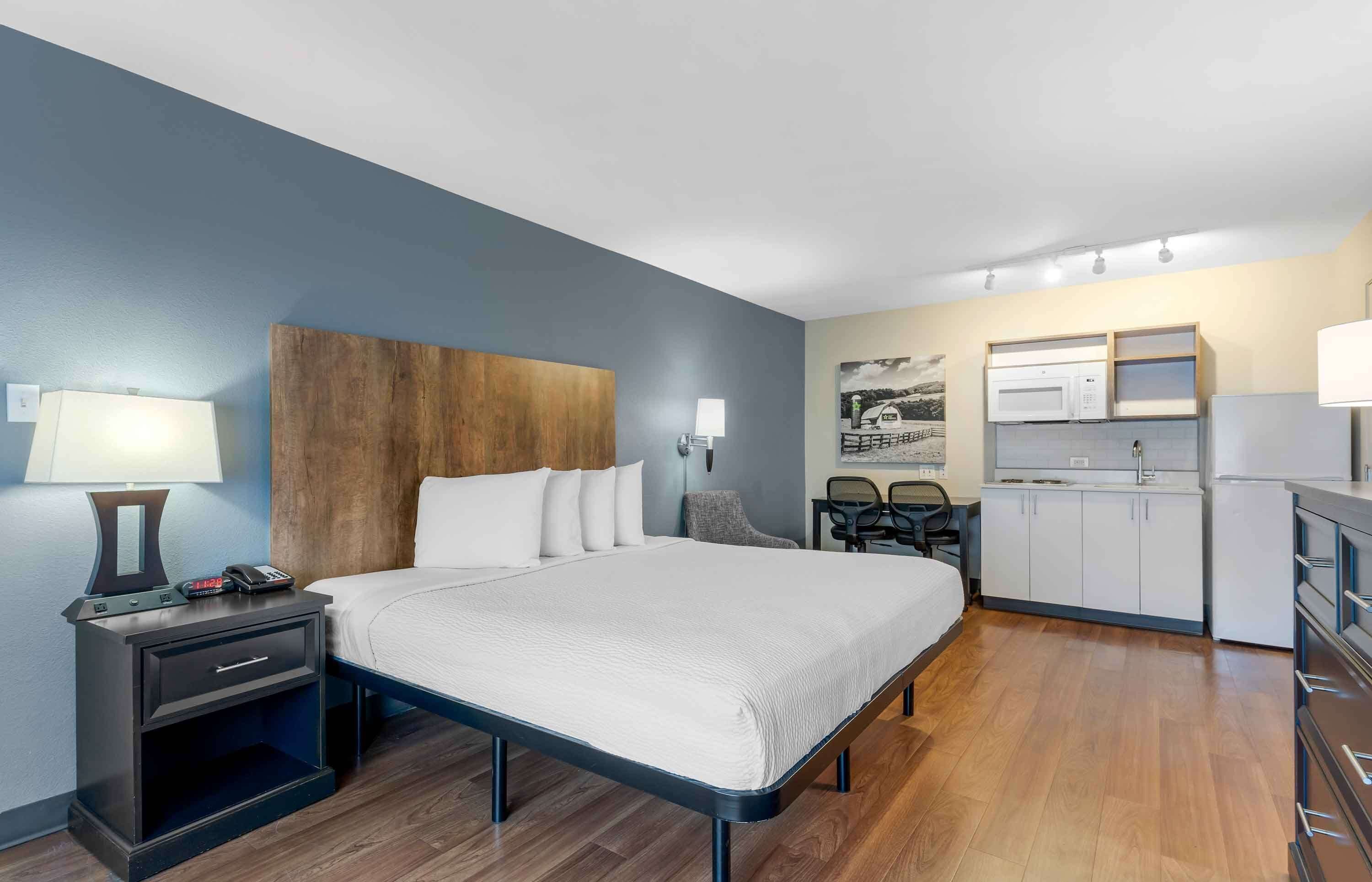 A hotel room with a bed, bedside table, lamp, phone, and a kitchenette with a microwave, refrigerator, and cabinets.