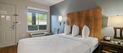 A hotel room with a large bed, a wooden headboard, a window with a view, a bedside table with a lamp, and a door.
