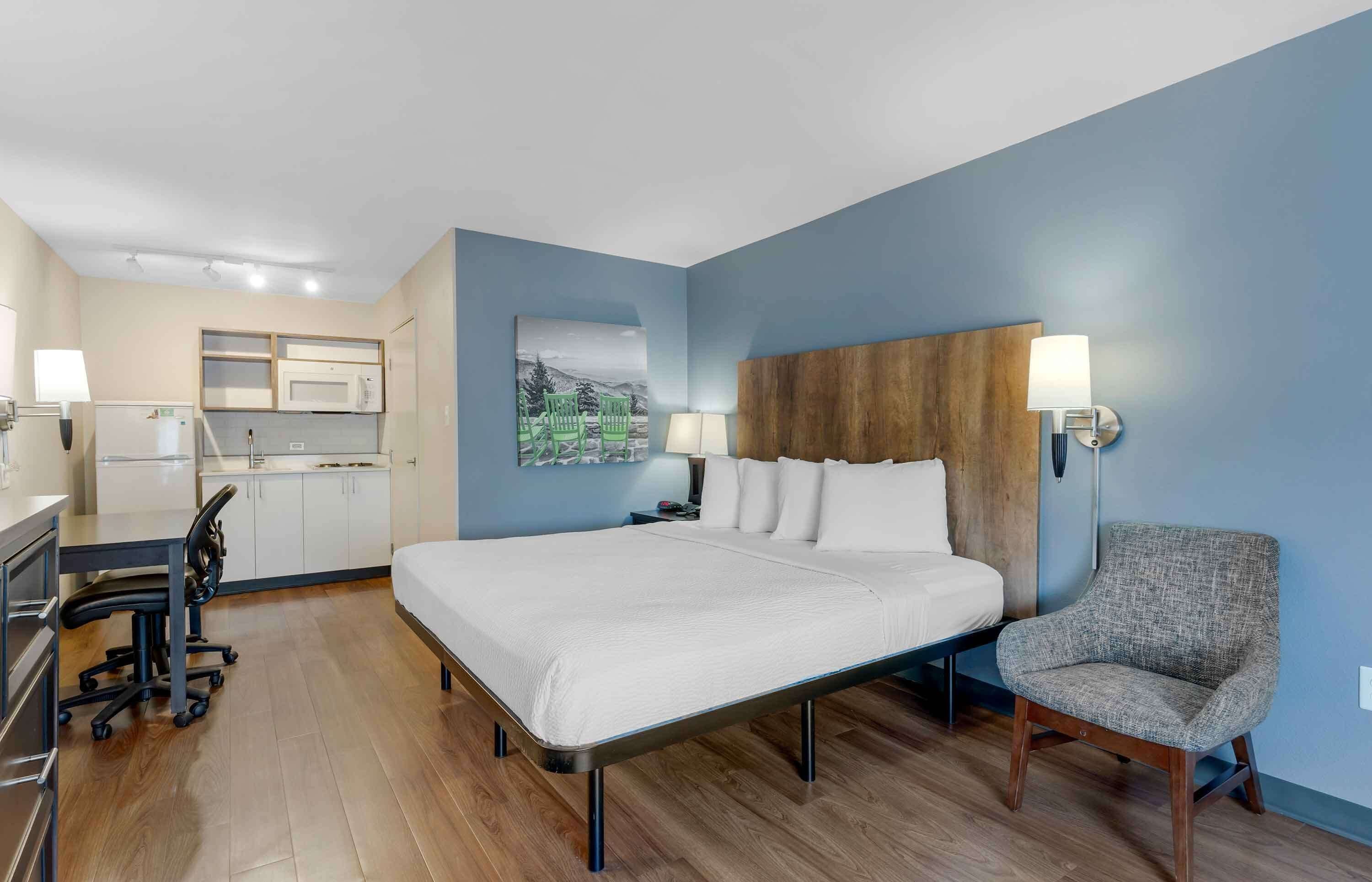 A hotel room with a bed, a chair, a desk, a kitchenette, and a painting on the wall.