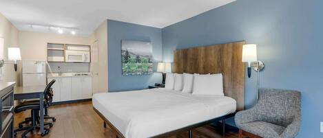 A hotel room with a bed, a chair, a desk, a kitchenette, and a painting on the wall.