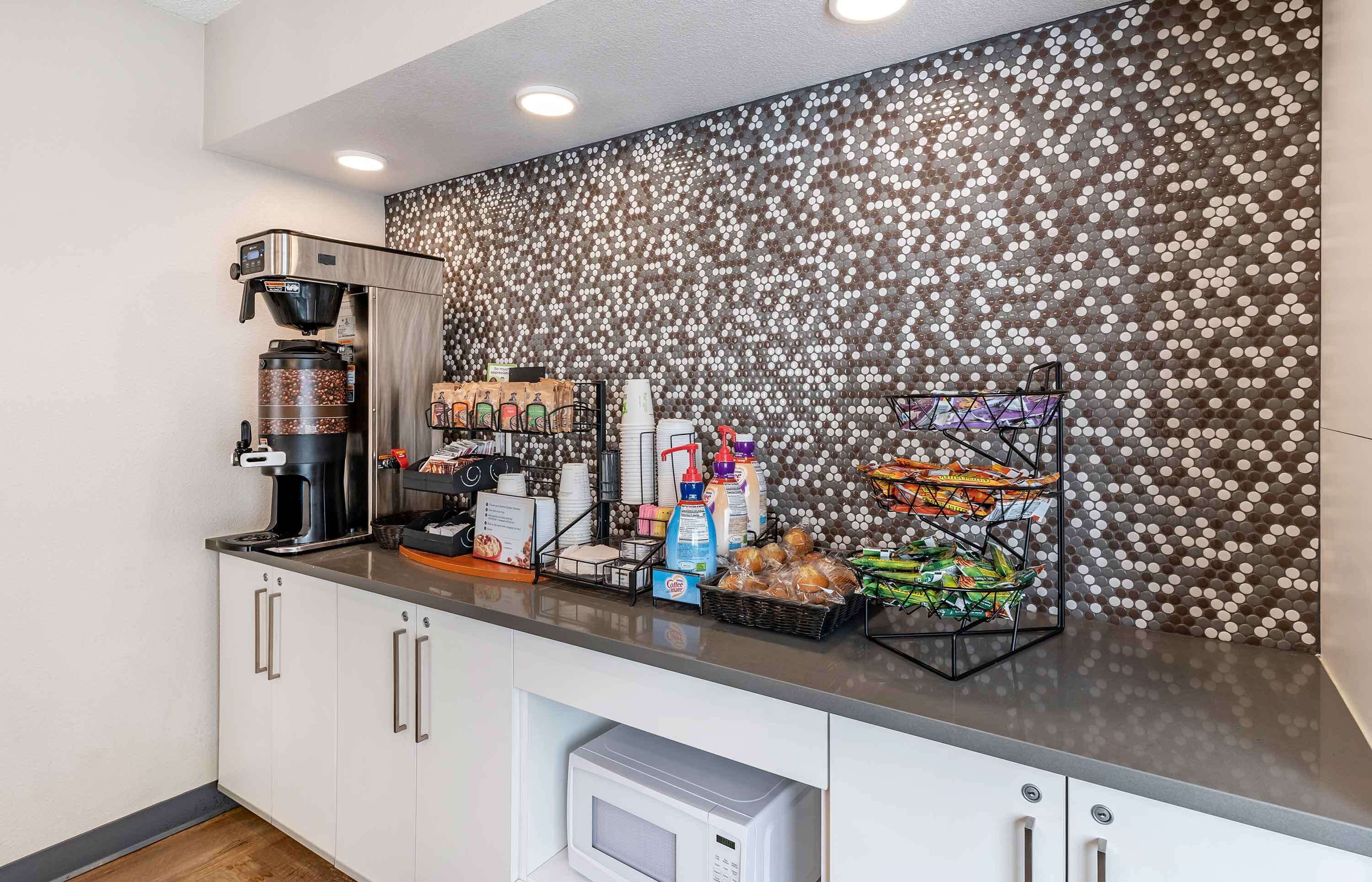 A modern kitchen area with a coffee machine, a microwave, and a beverage dispenser. There are various food items and condiments on the counter.