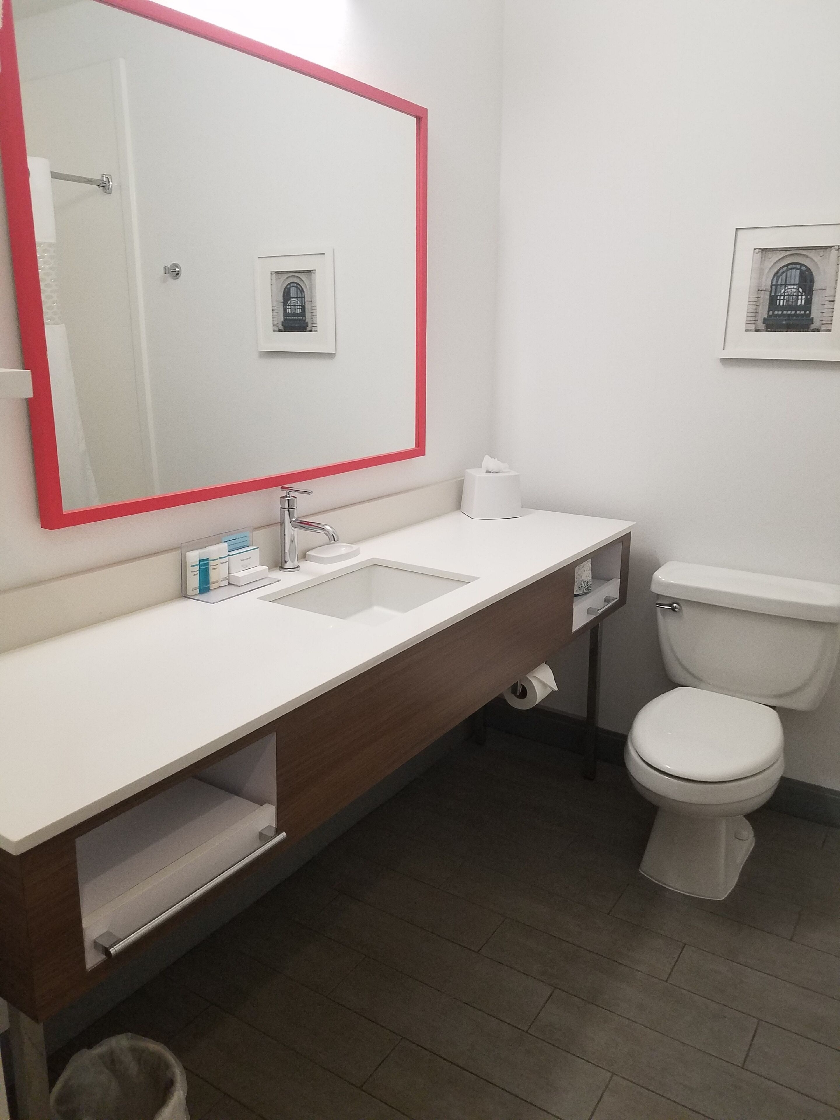 Un baño moderno con un lavamanos blanco, un espejo con marco rojo, un inodoro y una pequeña foto enmarcada en la pared.