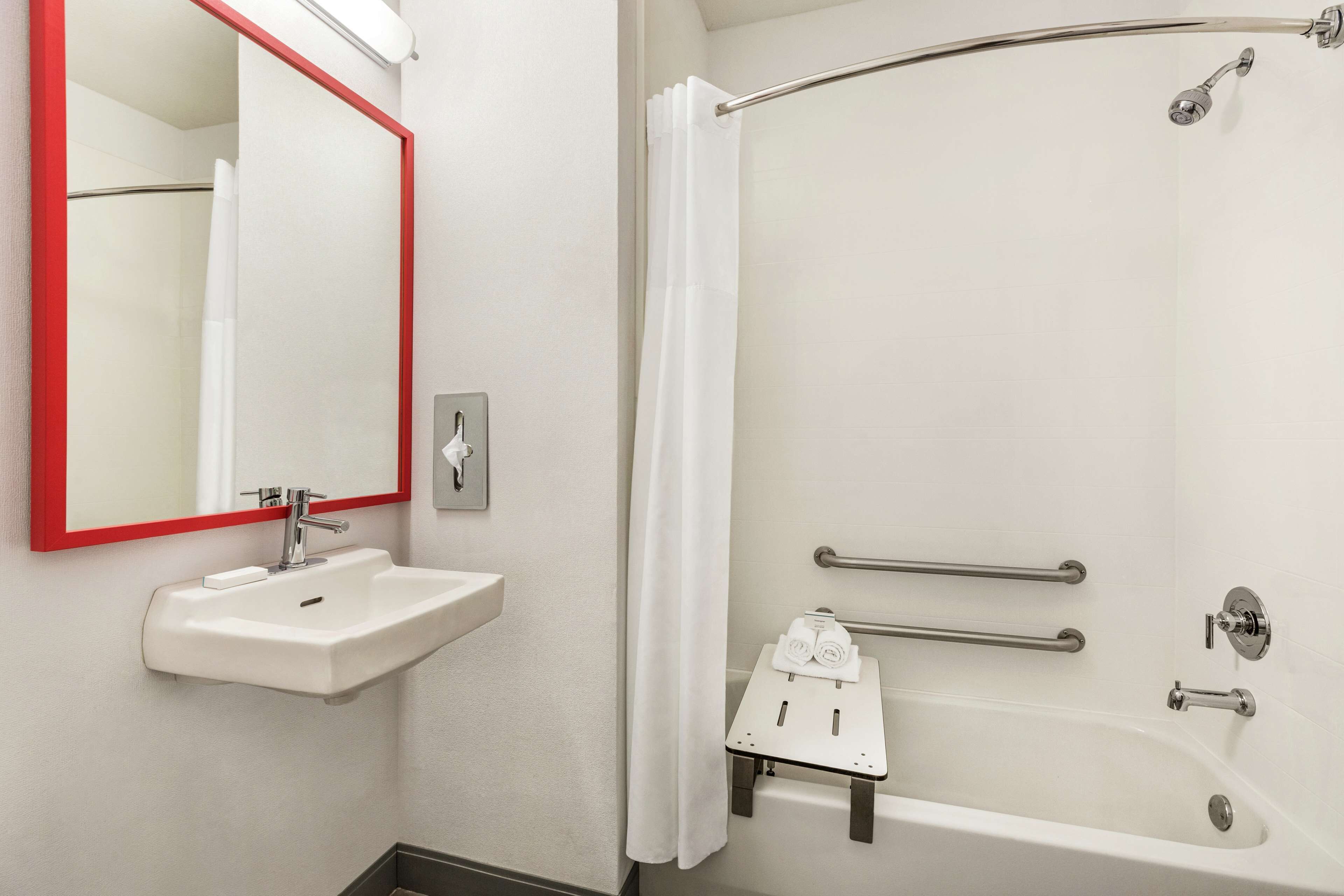A bathroom with a white sink, a mirror with a red frame, a shower with a curved glass door, and a small white shelf with towels.